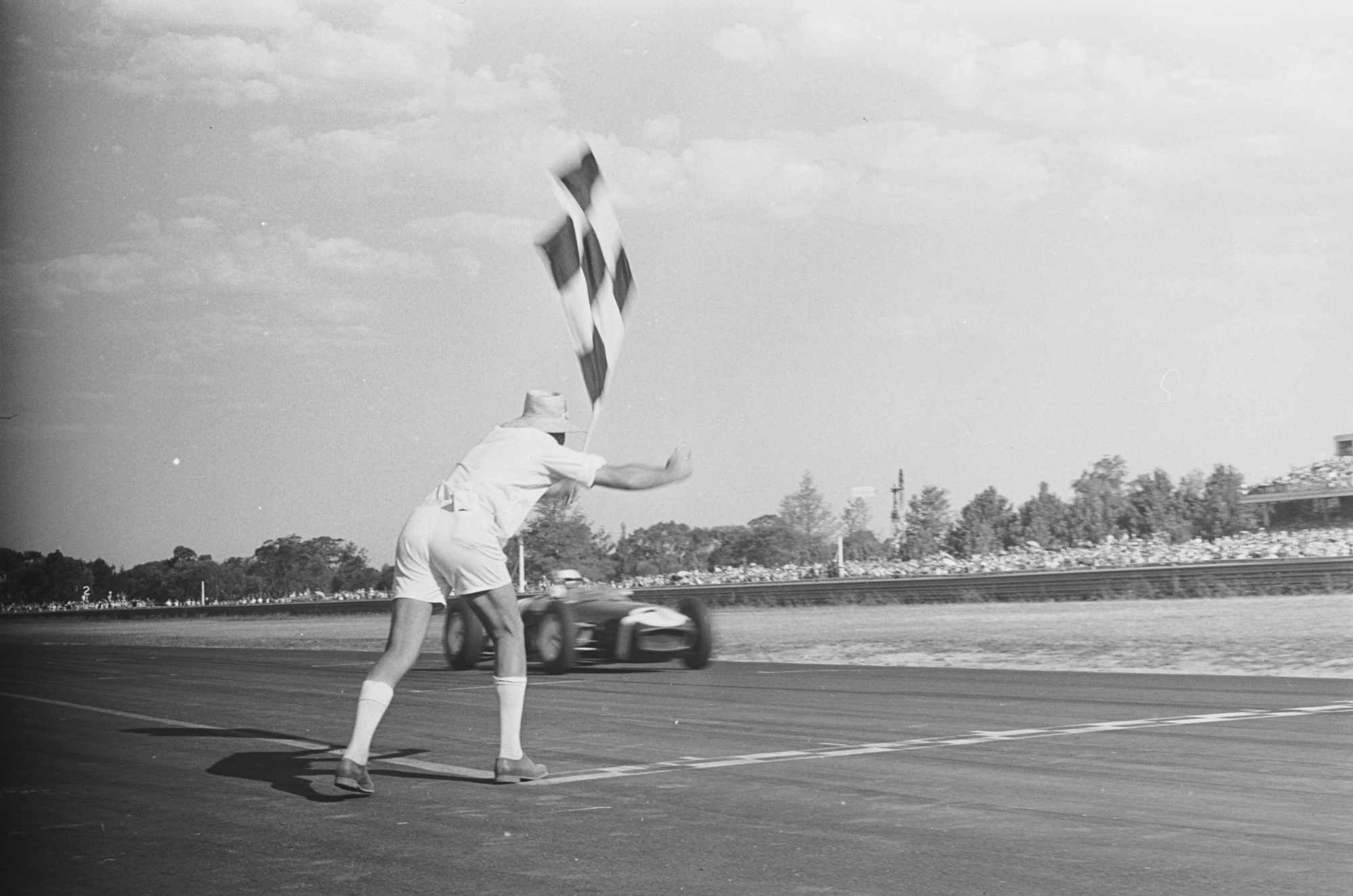 A race official enthusiasticaly waves the chequered flag as Stirling Moss crosses the line to win the inuagural Warwick Farm 100